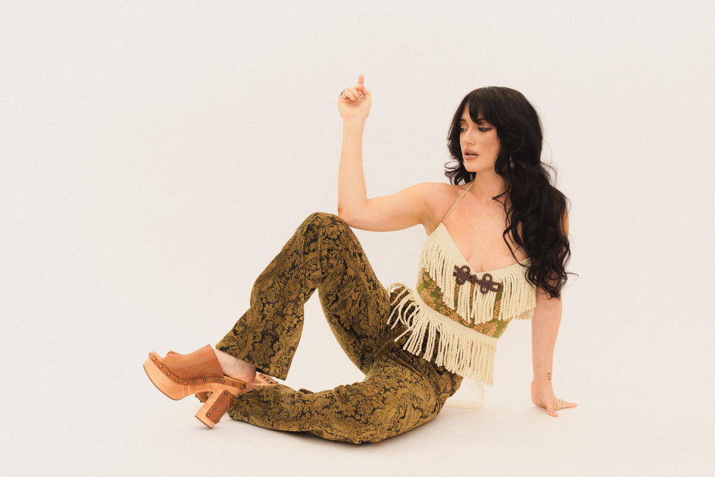Woman in a stylish outfit with a fringed top and patterned pants sitting on a white background