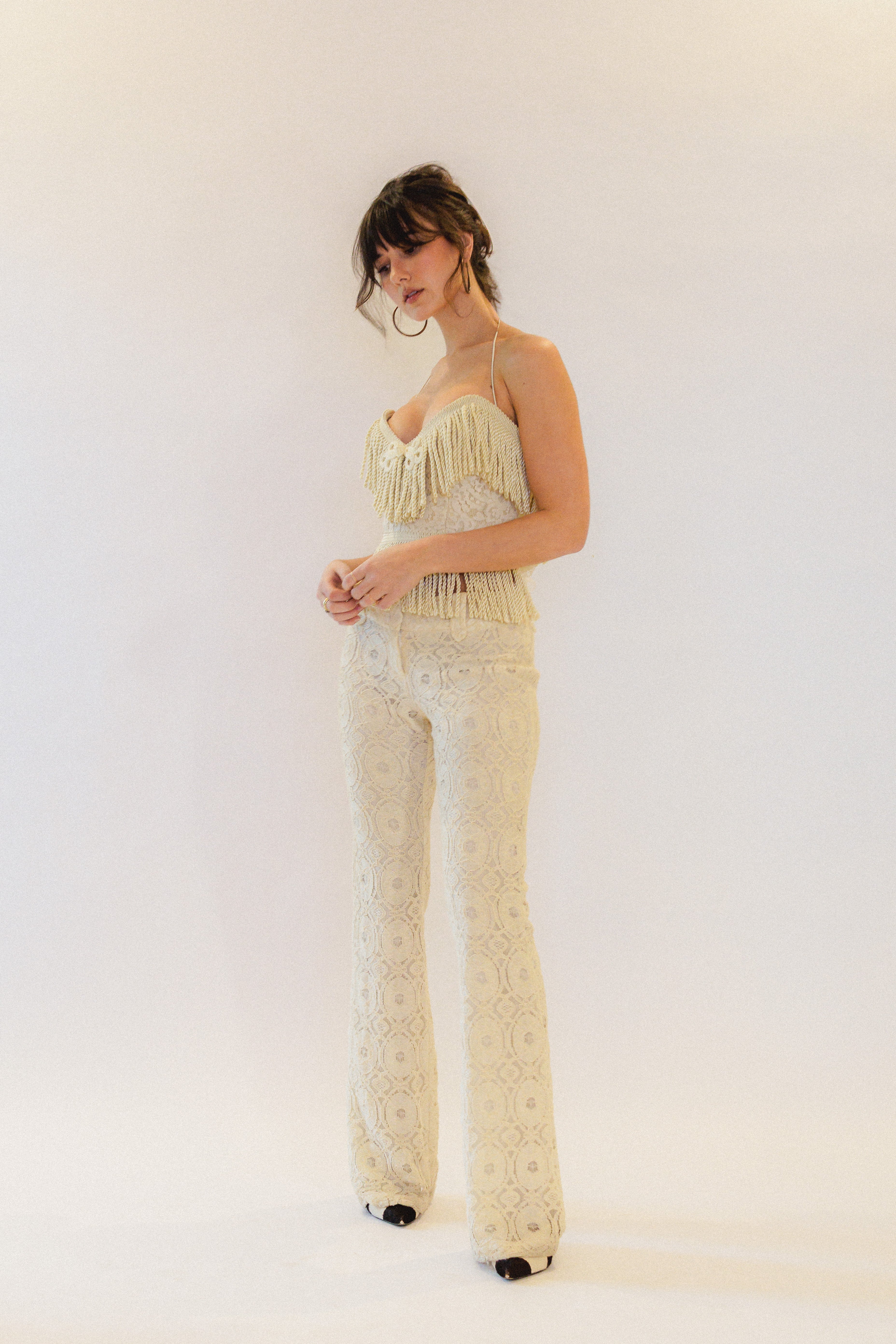 Woman wearing a white lace fringe corset top and pants against a plain background