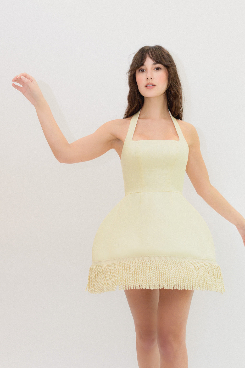 A woman modeling a pale yellow halter mini dress with a fringed hem against a plain light gray studio background.