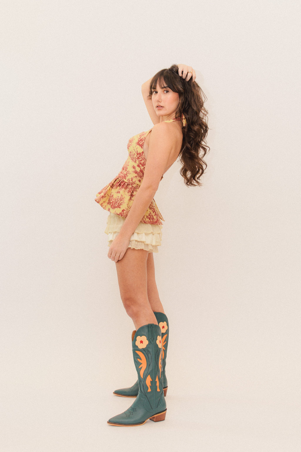 Woman wearing a red and gold halter peplum top and blue cowboy boots on a plain background