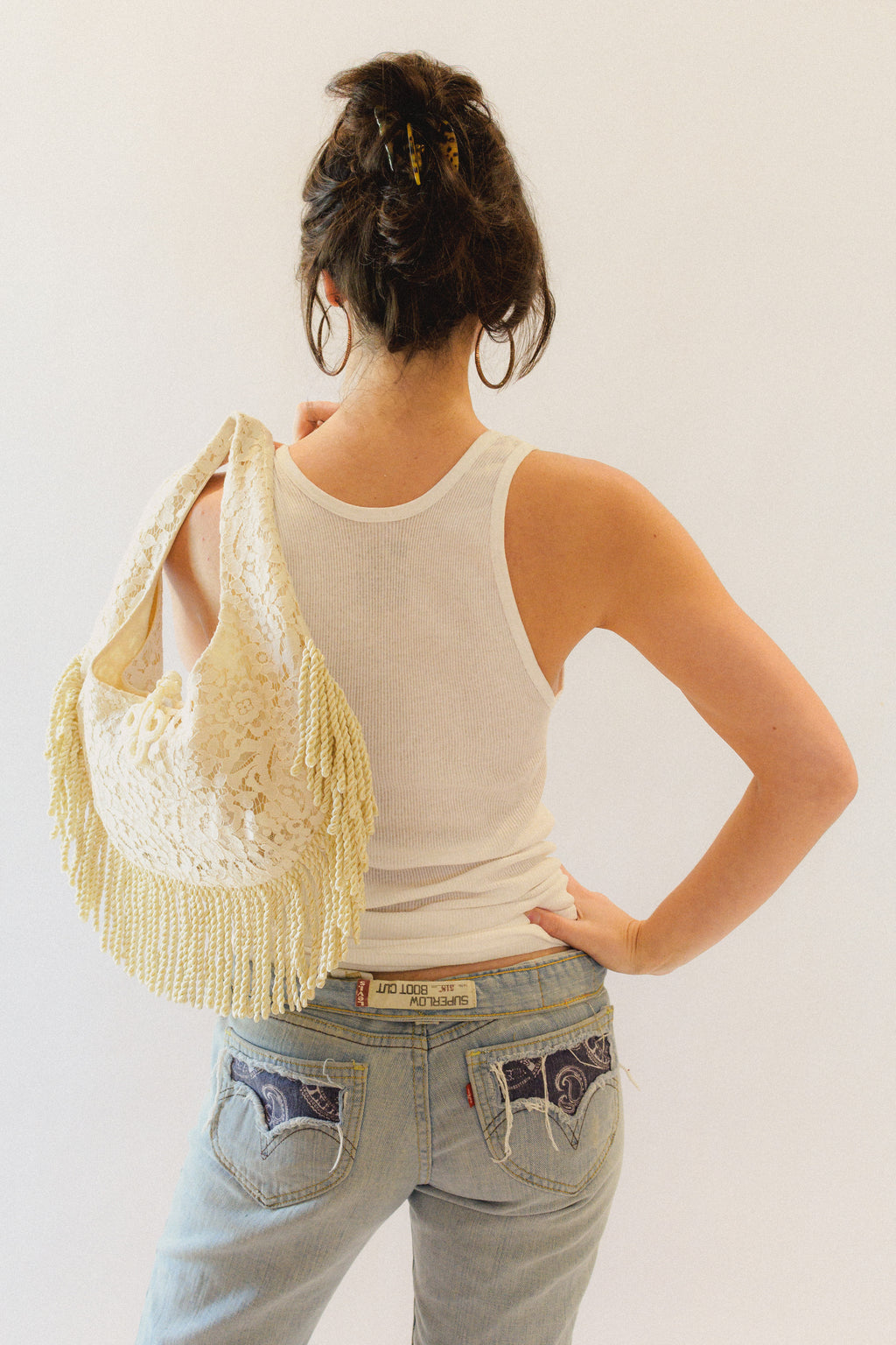 Person wearing a white tank top and light blue jeans with a cream lace bag with fringe over one shoulder.