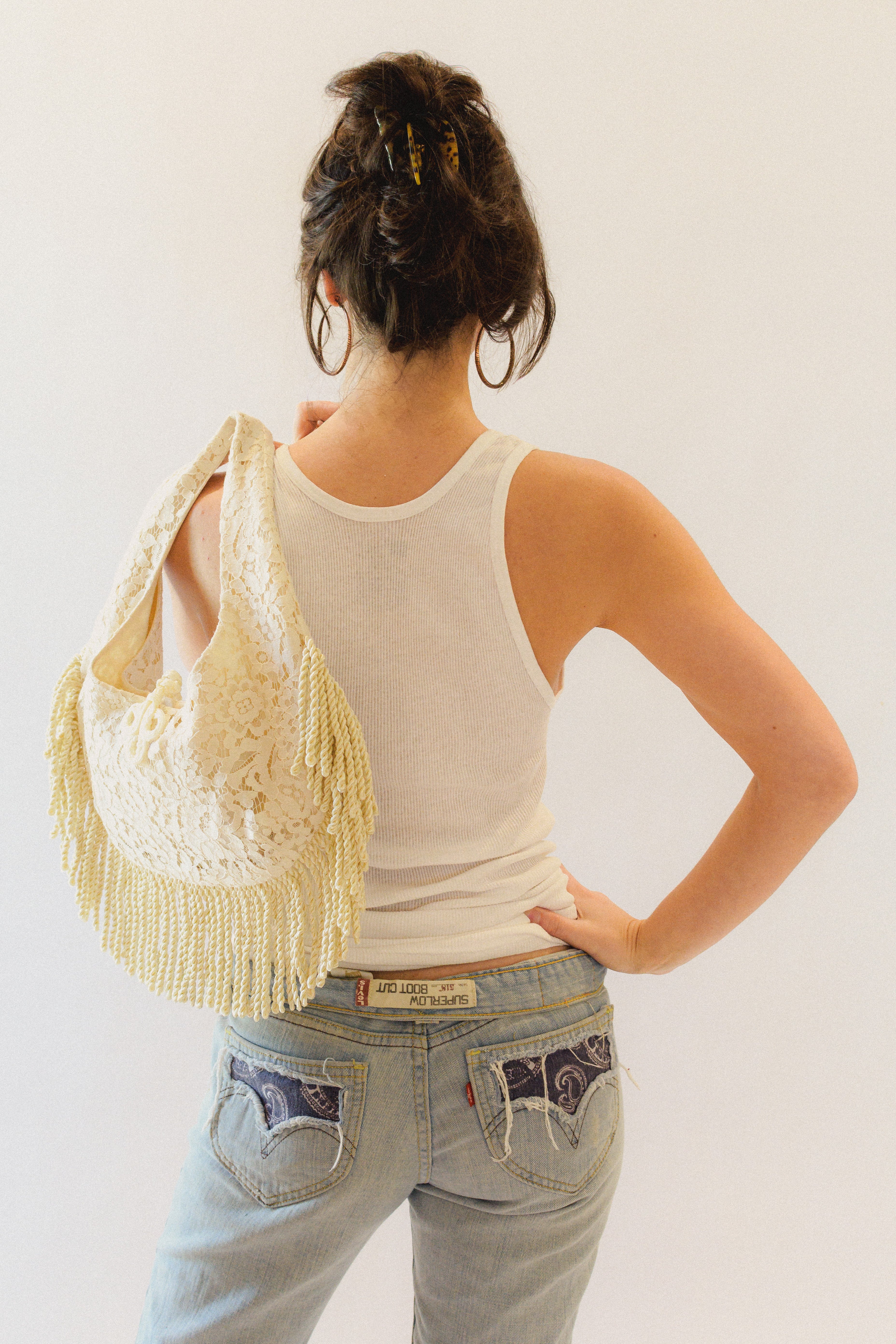 Person wearing a white tank top and light blue jeans with a cream lace bag with fringe over one shoulder.