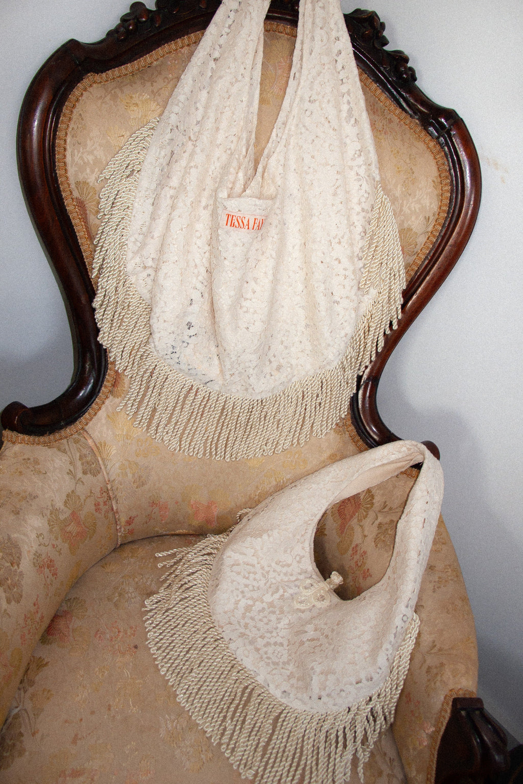 Two white handbags with fringes on a decorative chair against a plain wall.