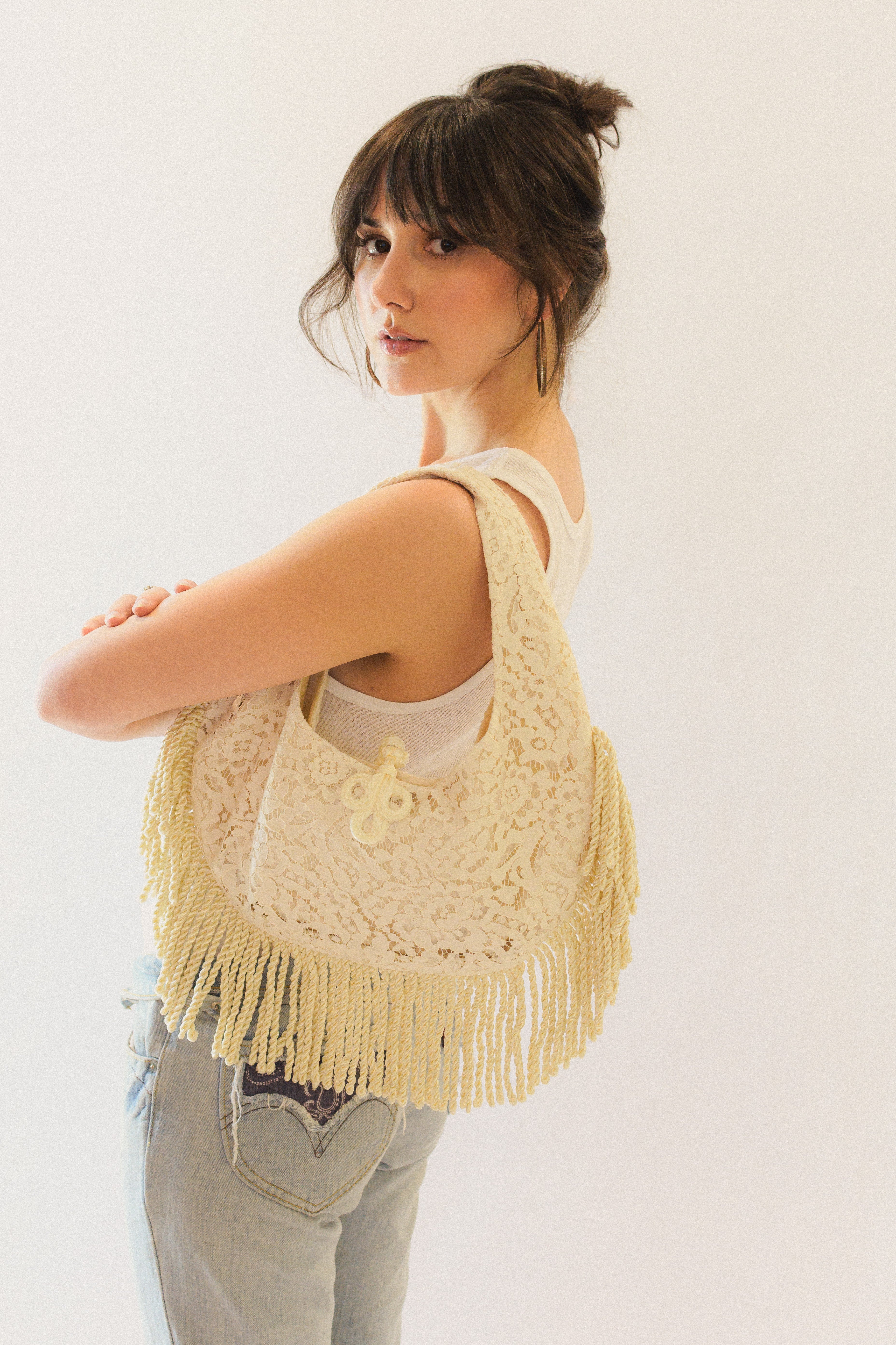 Woman wearing a lace shoulder bag with fringe and frog closure on a plain background