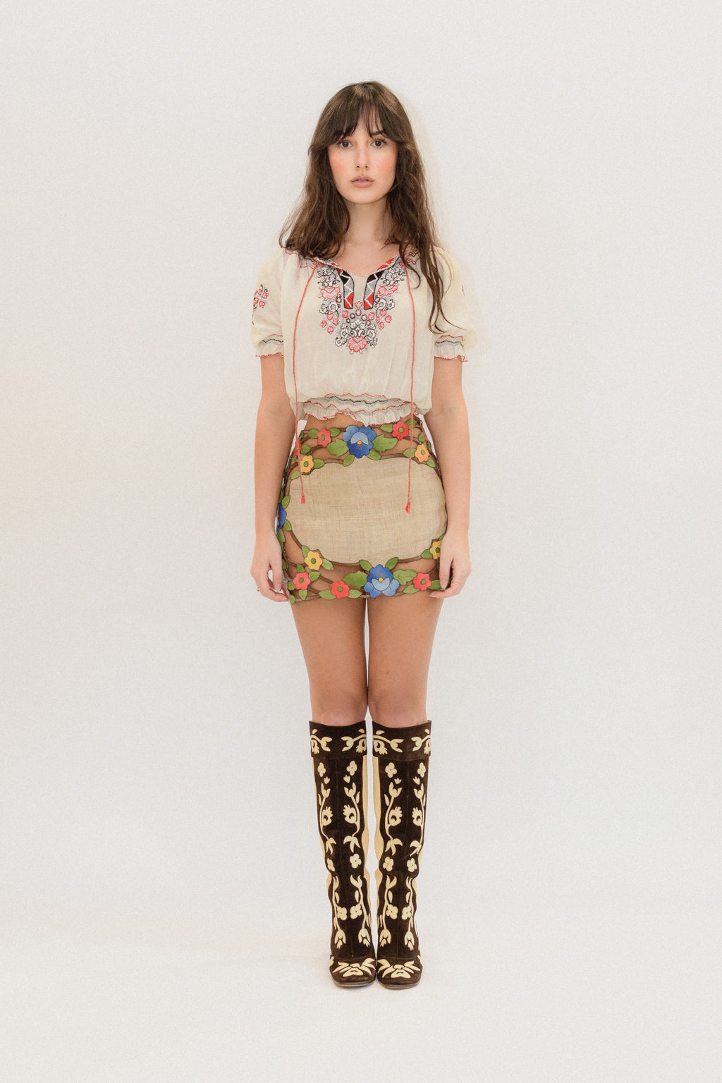 A model stands against a plain white studio background wearing a cream short-sleeve embroidered blouse, a beige mini skirt with colorful floral embroidery, and tall dark brown knee-high boots with gold patterns.
