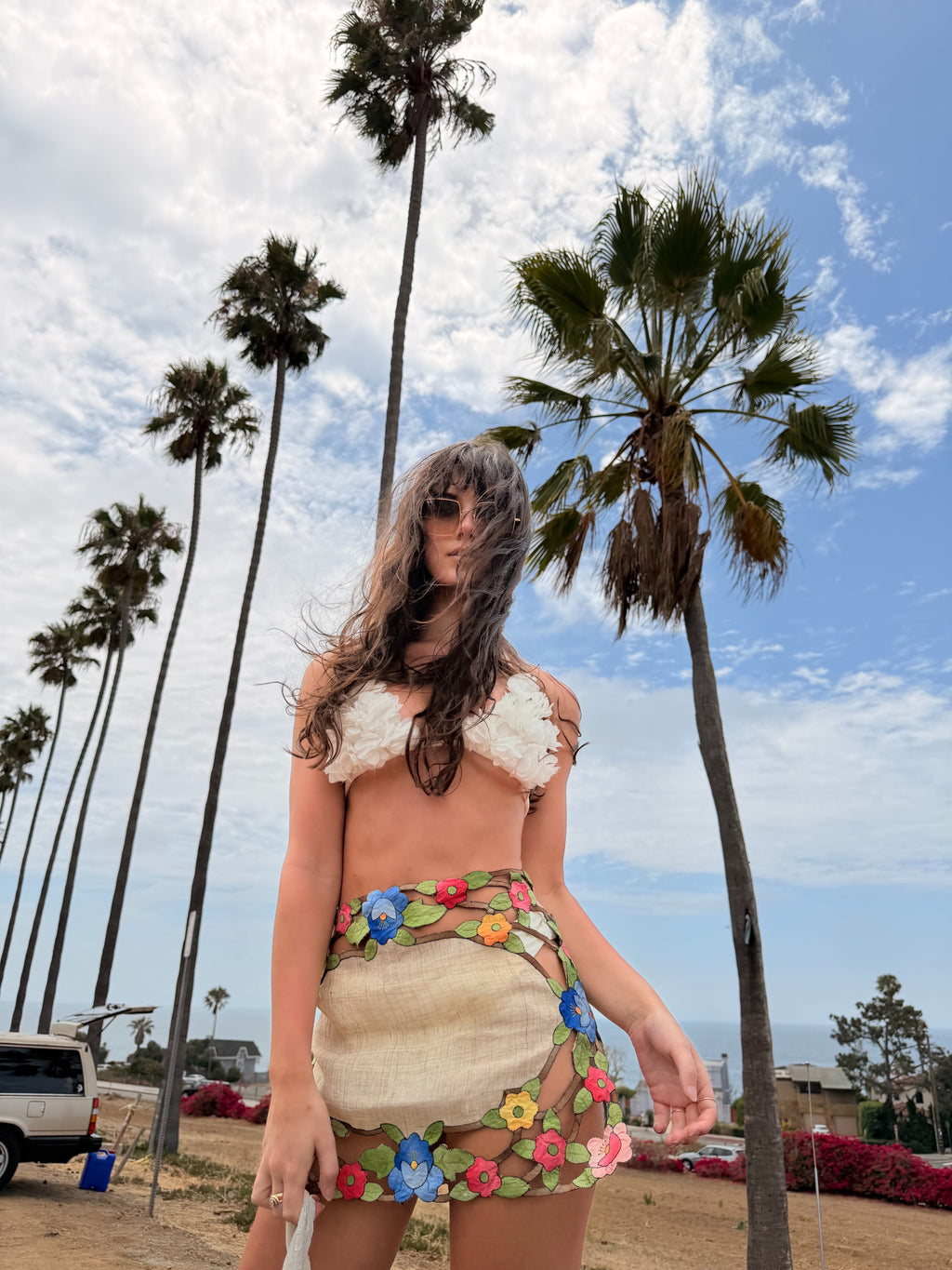 A woman stands outdoors on a beachside path with tall palm trees and blue sky behind her, wearing a textured cream bikini top and a matching floral mesh mini skirt with a green grass print waistband.