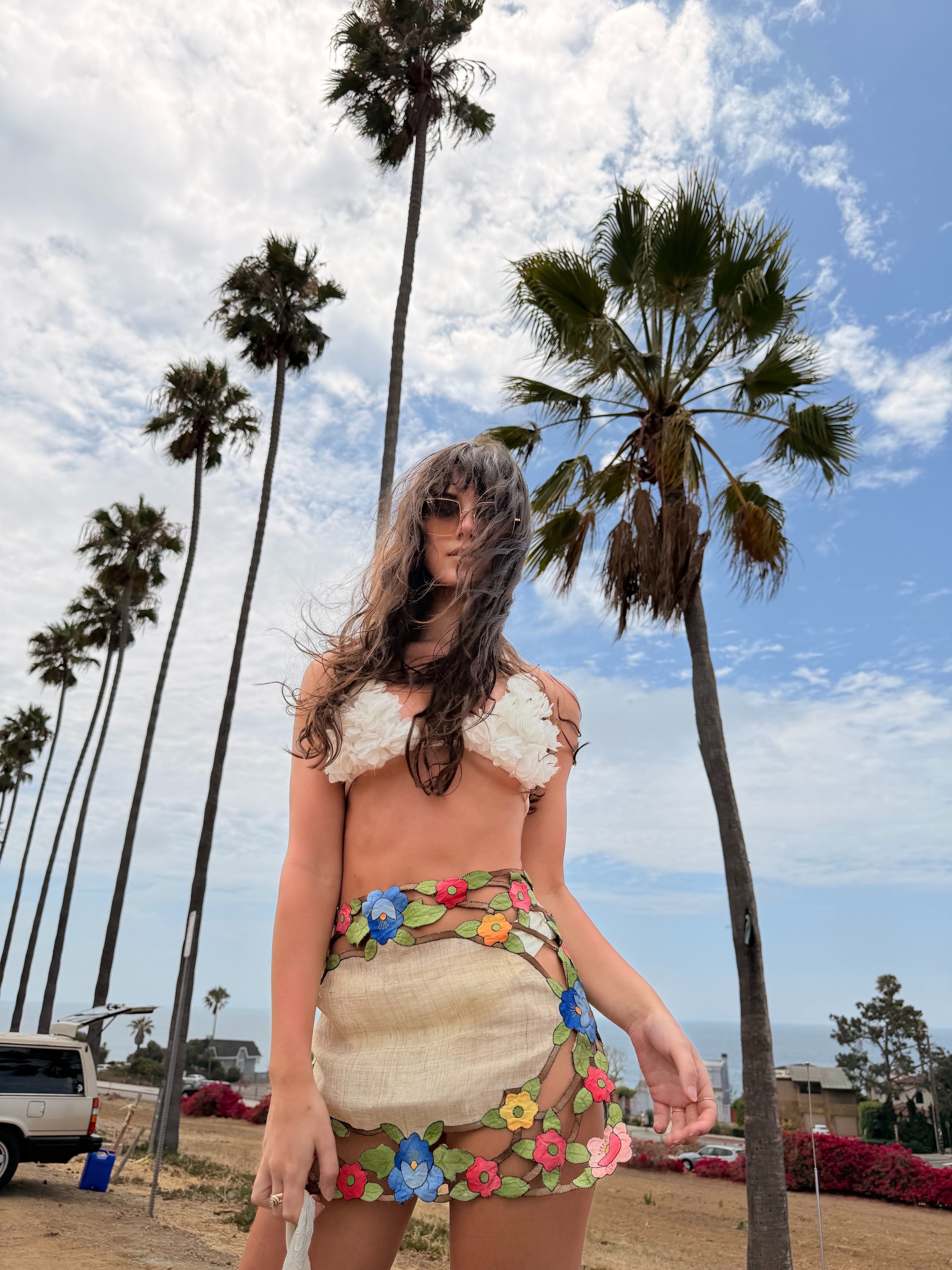 A woman stands outdoors on a beachside path with tall palm trees and blue sky behind her, wearing a textured cream bikini top and a matching floral mesh mini skirt with a green grass print waistband.