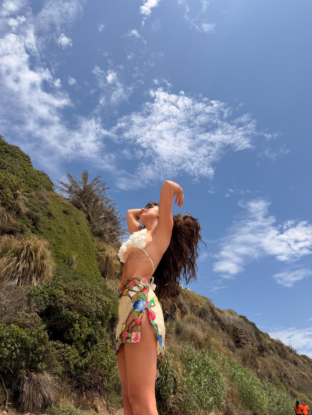 A woman posing outdoors against a blue sky with clouds, wearing a white ruffled bikini top and a beige floral bikini bottom.